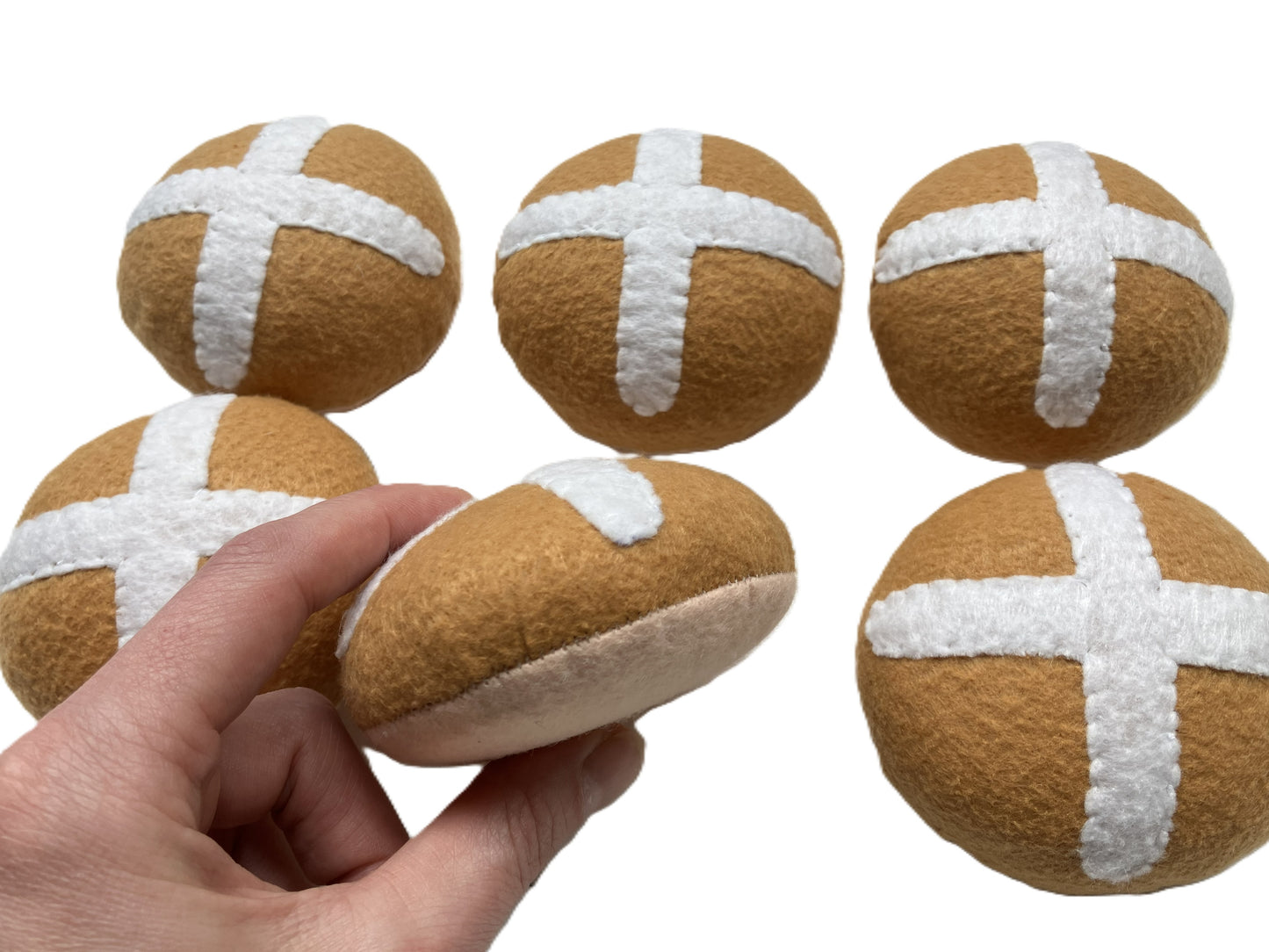 six brown and white cross-shaped felt hot cross buns with a hand holding one of them on a white background.