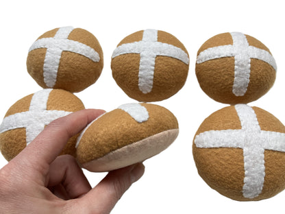 six brown and white cross-shaped felt hot cross buns with a hand holding one of them on a white background.