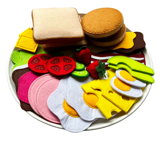 Colorful handmade felt food items presented on a plate including bread, pancakes, tomatoes, cheese, fried and boiled eggs, ham, spreads, avocados, cucumbers, strawberries and assorted spreads

