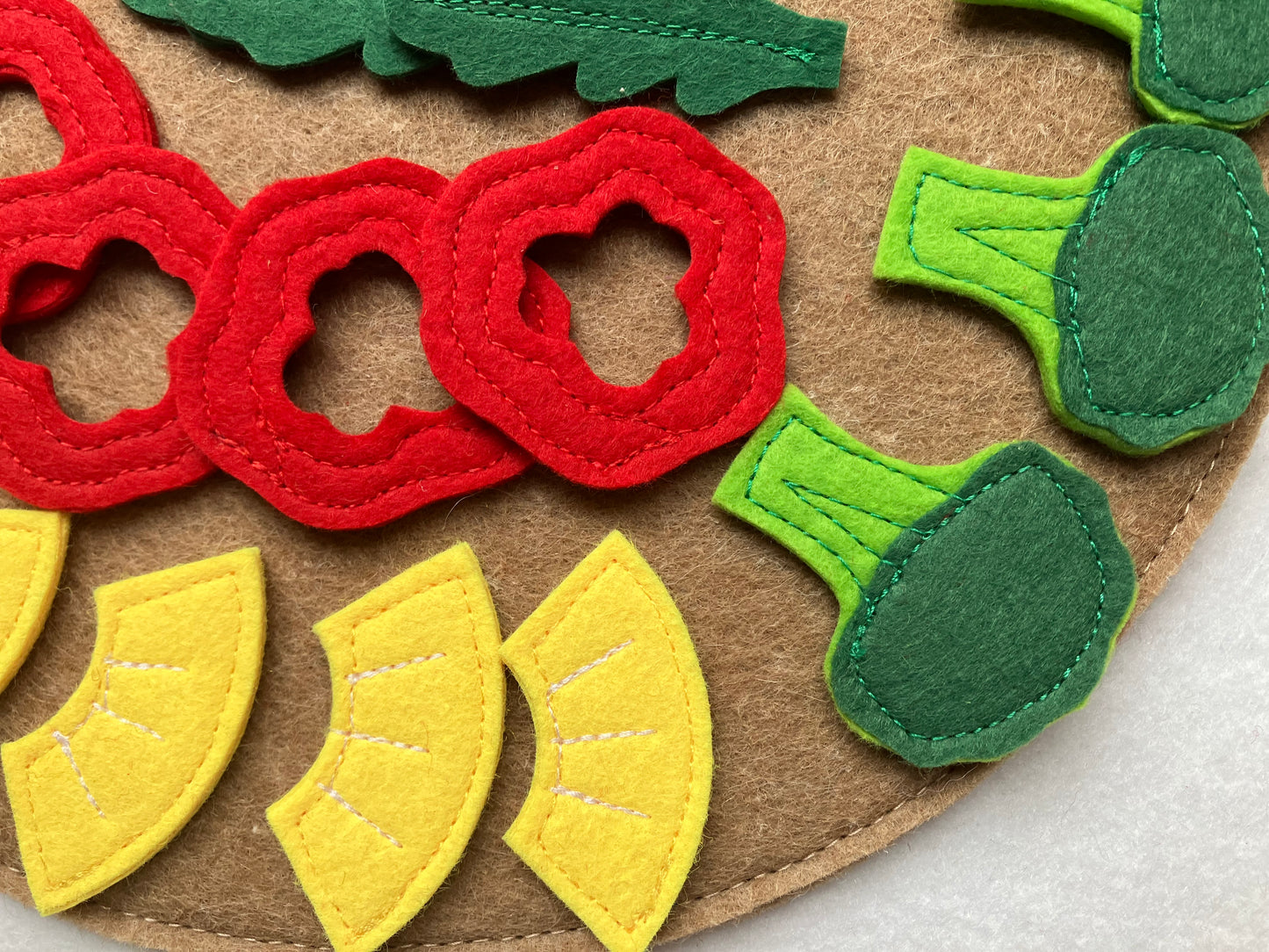 Farbige handgemachte Filz-Lebensmittel als Pizzabeläge: Ananas, Brokkoli, Pilze, Pepperoni und Paprika, arrangiert auf einem filzigen Untergrund vor braunem Hintergrund.
