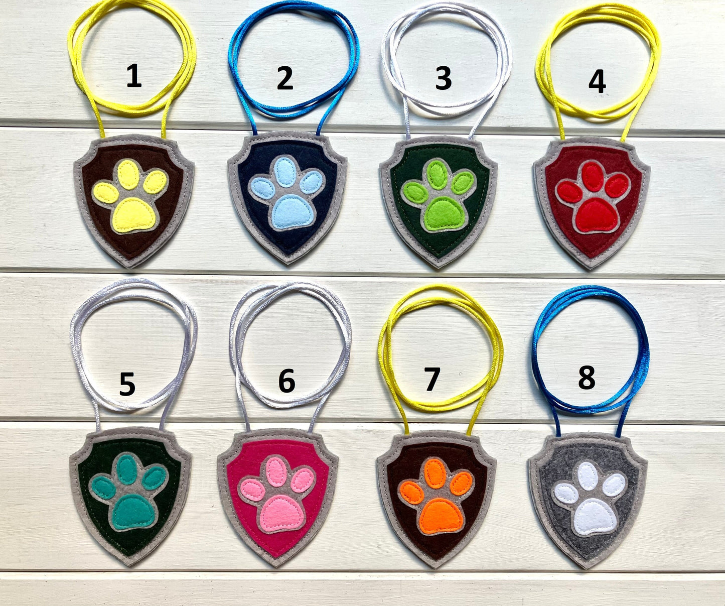 eight handmade shield-shaped  felt badges with hanging strings in various colors each representing a paw patrol character