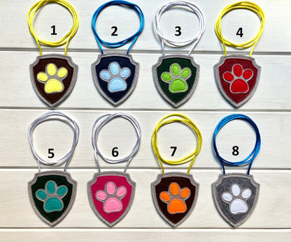 eight handmade shield-shaped  felt badges with hanging strings in various colors each representing a paw patrol character