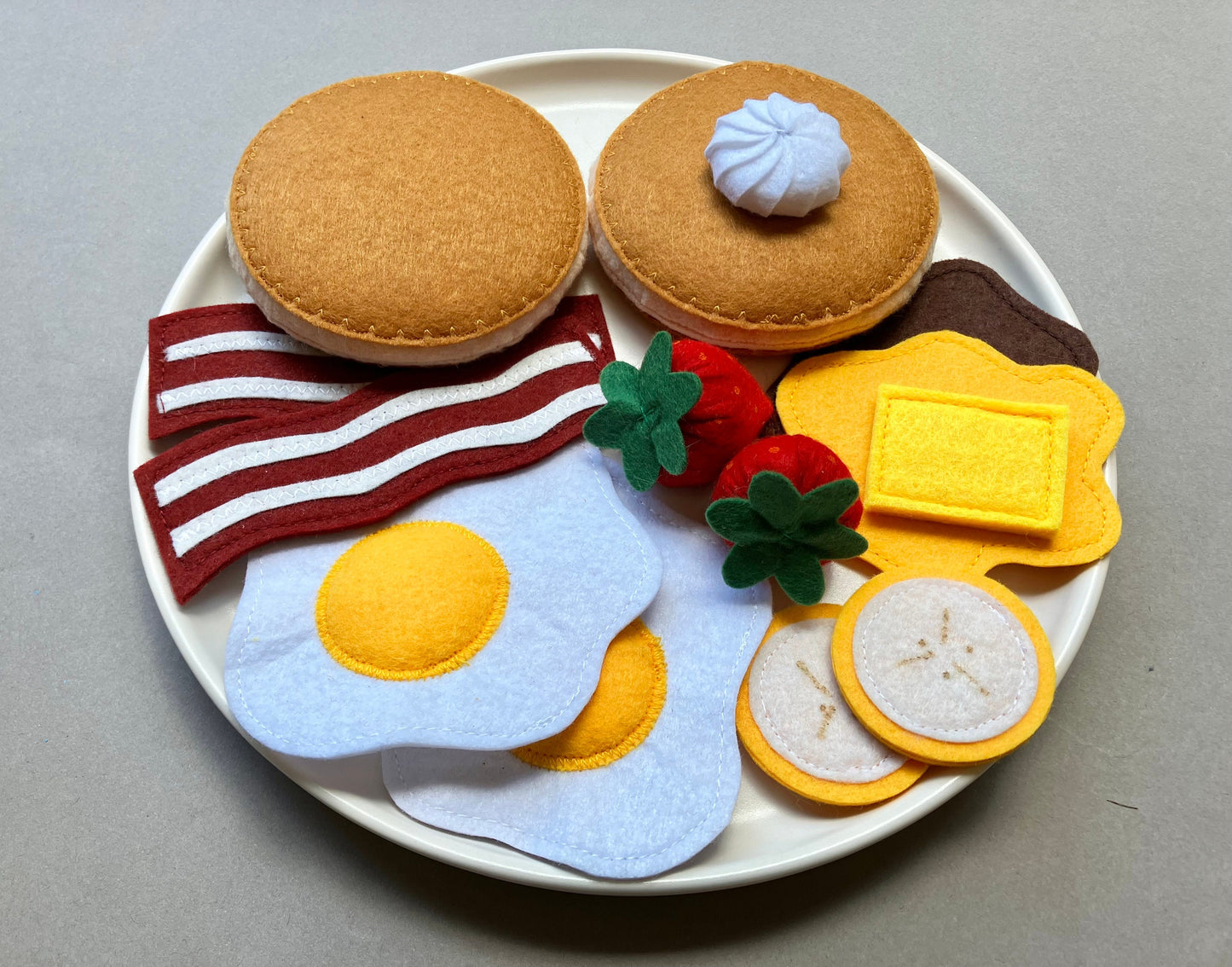Handmade Felt food set resembling a breakfast plate with pancakes eggs, bacon, bananas, spreads and berries on a gray background.