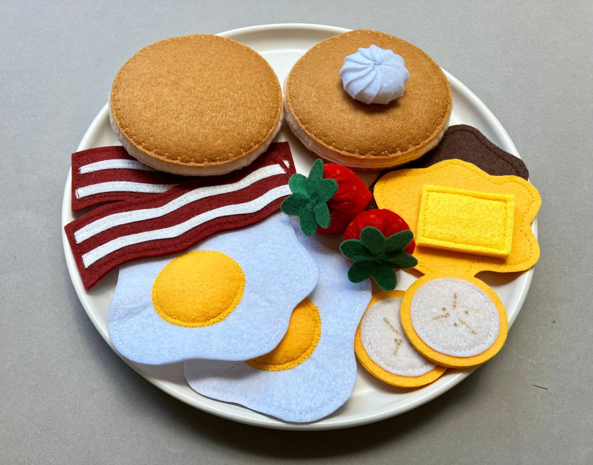 Handmade Felt food set resembling a breakfast plate with pancakes eggs, bacon, bananas, spreads and berries on a gray background.