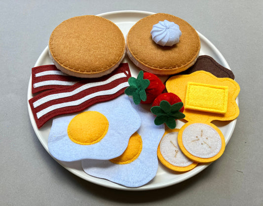 Handmade Felt food set resembling a breakfast plate with pancakes eggs, bacon, bananas, spreads and berries on a gray background.